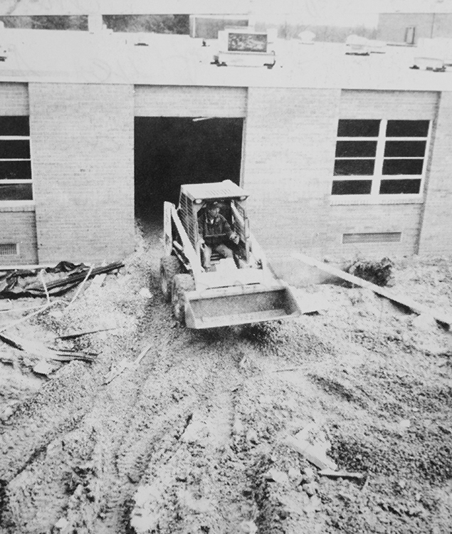 Black and white photograph of a construction worker operating a Bobcat next to the building.